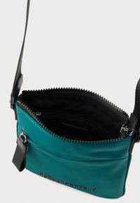 Tyrkysová kabelka cez rameno s čiernym zipsom a remienkom, s kontrastným čiernym vnútrom a vnútornou vreckom so zipsom pre lepšiu organizáciu.