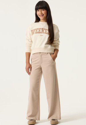 Teenager-Mädchen mit langen dunklen Haaren, das einen beigen "WEEKEND"-Sweatshirt, weite beige Nadelstreifenhose und beige Turnschuhe trägt, steht mit einer Hand in der Tasche.