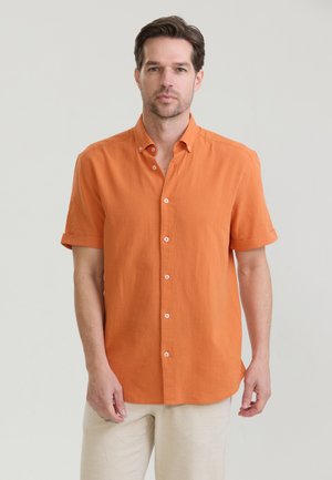 Mann mit kurzen braunen Haaren, der ein orangefarbenes kurzärmeliges Hemd mit Knöpfen und helle Hosen trägt, steht vor einem einfarbigen Hintergrund.