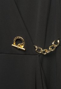 Svart tygplagg med ett guld cirkelfäste med en horisontell stång och ett guld kedjedetalj längs sömmen.