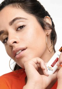 Flacon de sérum blanc avec des accents orange, étiqueté "Revitalift Clinical", tenu par une personne au teint clair, portant un haut orange vif.