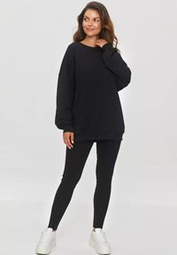 Czarna oversize'owa bluza z miękkiego materiału, zestawiona z czarnymi legginsami i białymi sneakersami. Posiada długie rękawy i okrągły dekolt.