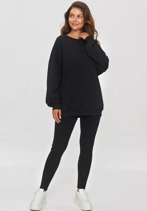 Czarna oversize'owa bluza z miękkiego materiału, zestawiona z czarnymi legginsami i białymi sneakersami. Posiada długie rękawy i okrągły dekolt.