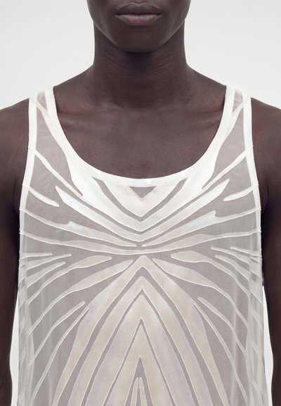Gros plan d'une personne portant un haut blanc sans manches, transparent, avec un motif géométrique et abstrait à l'avant.