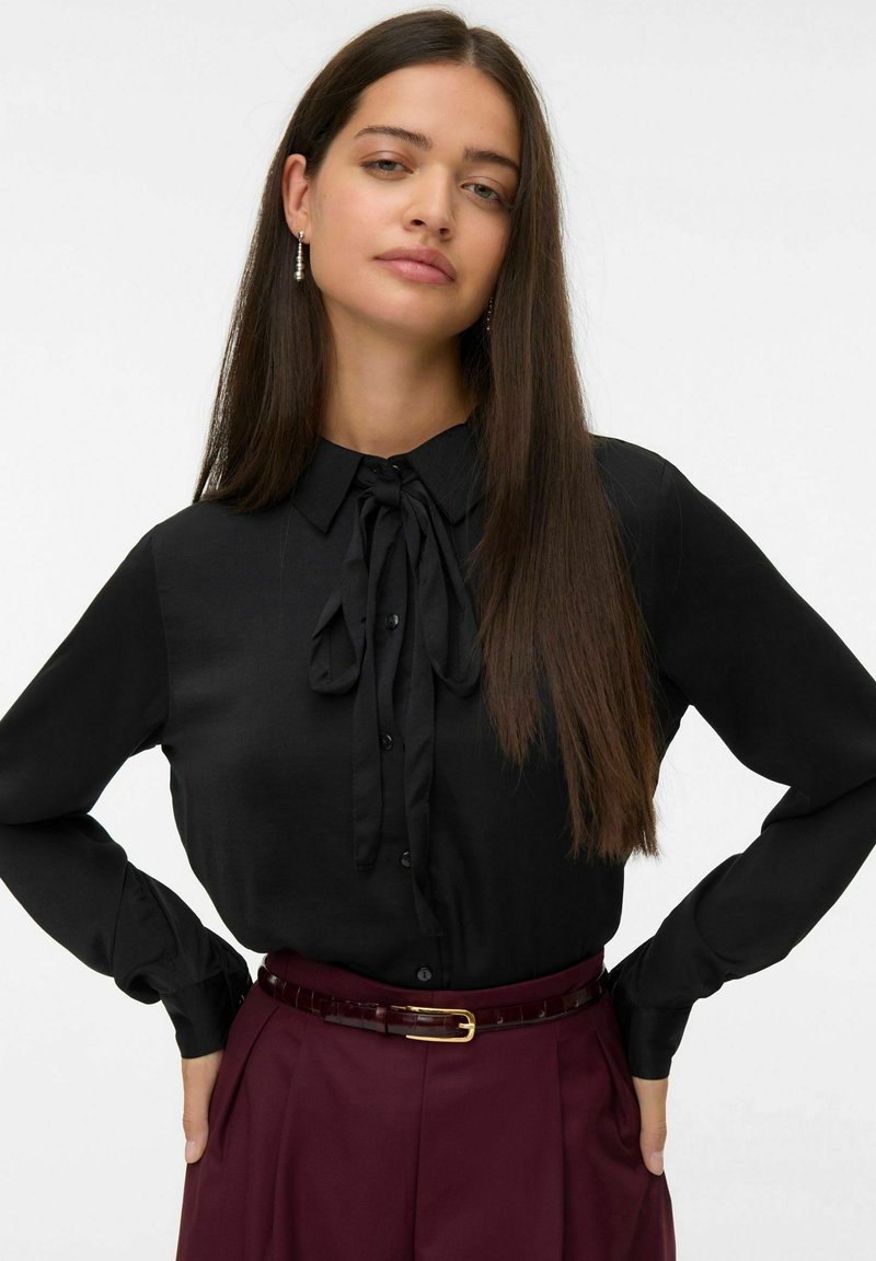 Zwarte blouse met een strik-kraag, gesloten met knopen aan de voorkant en lange mouwen. Gekleed met hooggeknoopte mahoniekleurige broek en een dunne bruine riem.