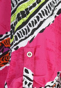 Farbiger Stoff mit rosa Hintergrund, der schwarze, weiße, orange und grüne Muster zeigt. Ein Knopfdetail ist am Rand sichtbar.