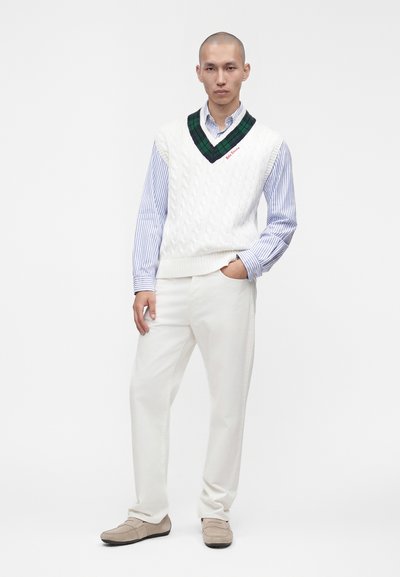 Homme portant un gilet en tricot torsadé blanc avec une bordure en V verte et noire, sur une chemise rayée bleue, un pantalon blanc et des mocassins beige.