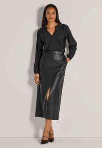 Schwarze Bluse mit langen Ärmeln und V-Ausschnitt, kombiniert mit einem figurbetonten schwarzen Lederrock mit einem Schlitz vorne. Abgerundet mit High Heels.