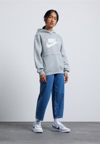 Pilkas džemperis su dideliu baltu Nike logotipu, priekinė kengūros kišenė, derinamas su plačiomis mėlynomis džinsomis ir baltomis sportbatėmis. Paprastas, kasdienis dizainas.