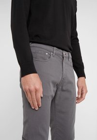 Jean en denim gris à coupe droite, avec braguette zippée, cinq poches et détails de couture subtils. Associé à un pull noir.