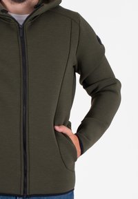 Felpa con zip di colore verde oliva, realizzata in un tessuto morbido e strutturato, dotata di cappuccio, tasche laterali e zipper nera, con un design aderente.