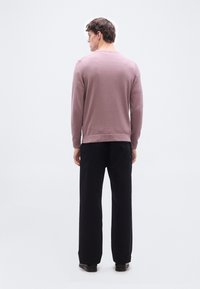 Άντρας που στέκεται με πλάτη φοράει μοβ πουλόβερ με μακριά μανίκια, μαύρο παντελόνι και μαύρα παπούτσια σε απλό λευκό φόντο.