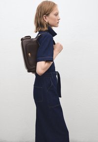 Hnedý kožený batoh s obdĺžnikovým tvarom a zlatými kovaniami, nosený cez tmavomodrý denimový overal s krátkymi rukávmi a opaskom v páse.