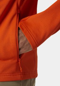 Pomarańczowa bluzka z długim rękawem o gładkiej fakturze. Posiada boczną kieszeń na zamek oraz regulowane mankiety, co podkreśla funkcjonalność i design.