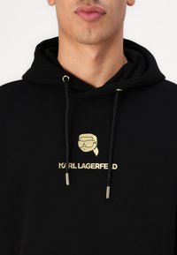 Felpa nera con cappuccio realizzata in tessuto morbido, caratterizzata da un logo grafico dorato e dal testo "KARL LAGERFELD". Corda regolabile con dettagli in oro.