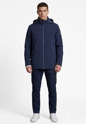 Hombre de pie vistiendo una chaqueta con capucha azul marino, pantalones a juego y zapatillas blancas sobre un fondo blanco liso.