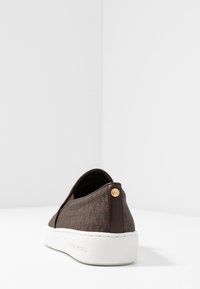 Baskets en textile à motif marron avec une semelle en caoutchouc blanche, présentant un accent en cuir au talon et un détail de logo doré.