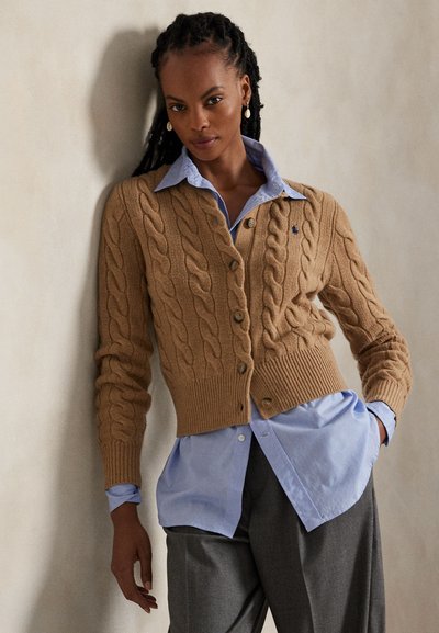 Femeie cu cozi lungi, purtând un cardigan de cașmir camel cu model torsadat peste o cămașă albastră și pantaloni gri, sprijinită de un perete neutru.