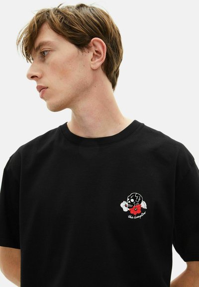 Camiseta de algodón negra con un pequeño diseño bordado de un cráneo con pétalos rojos y el texto "the longer." Cuello de barco y mangas cortas.