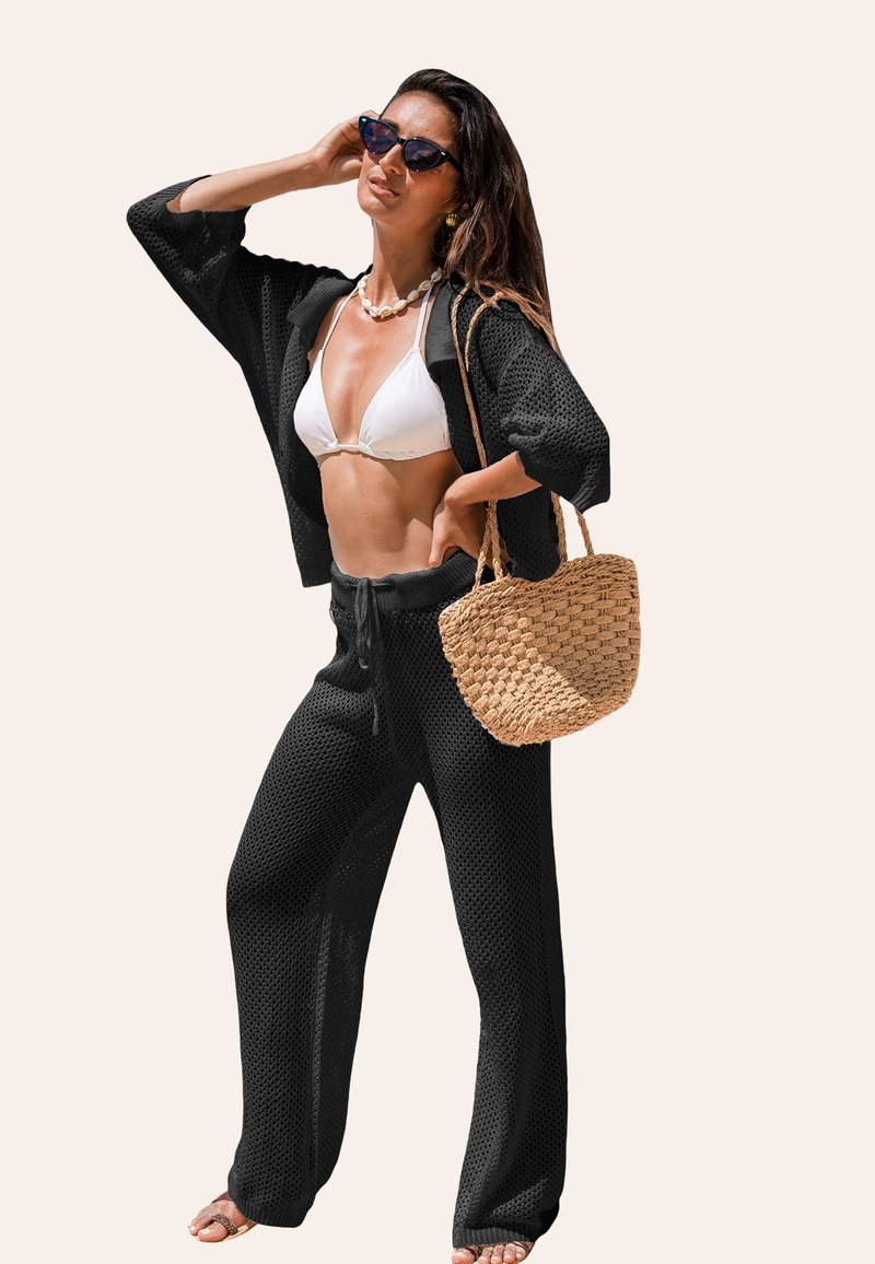 Cardigan nero a maglia aperta sopra un costume da bagno bianco, abbinato a pantaloni neri con coulisse. Accessoriato con una borsa in paglia intrecciata e occhiali da sole.