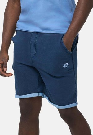 Homme portant un short bleu foncé à ourlet roulé avec des poches et une chemise bleu clair, debout devant un fond uni.