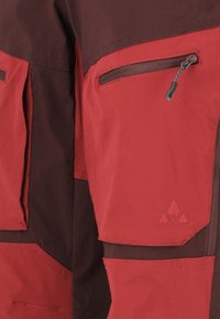 Pantalons d'extérieur rouges et bruns avec des poches zippées, en tissu robuste, avec des coutures renforcées et un accent logo en bas à droite.
