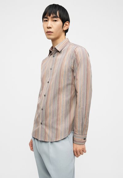 Νέος άνδρας που φοράει πουκάμισο με μακριά μανίκια και πολύχρωμες ρίγες, κουμπωτό, και ανοιχτόχρωμο γκρι παντελόνι, εναντίον ενός απλού λευκού φόντου.