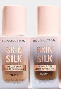 To flasker med Revolution Skin Silk foundation i rosa emballasje med rosagull aksenter, merket F12.7NC og F14.9NC, 23ml hver.