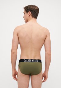 Maslinaste zelene kupaće gaće s crnim pojasom na kojem je bijeli natpis "CALVIN KLEIN". Glatka tekstura, prilagođen dizajn i nizak struk.