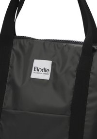 Svart nylon tote-väska med textilaxelremmar, med en vit etikett som har texten "Elodie STOCKHOLM SWEDEN" och dragkedjeöppning.