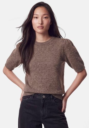 Femme aux longs cheveux foncés portant un pull en maille marron à manches courtes et un jean noir taille haute, debout les mains sur les hanches.
