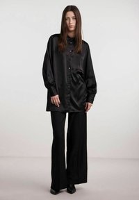 Black satin oversized shirt with button closure, long sleeves, and a glossy texture, paired with wide-leg black trousers and pointed heels.