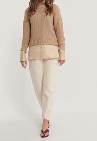 Beige strukturierter Strickpullover über einem hellbeigen Hemd, kombiniert mit locker sitzenden cremefarbenen Hosen und dunklen Sandalen.