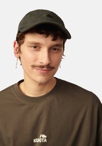 Ciemnozielona czapka baseballowa z haftowanym logo, z zakrzywionym wizjerem. Model nosi brązowy T-shirt z białym logo i srebrne kolczyki.