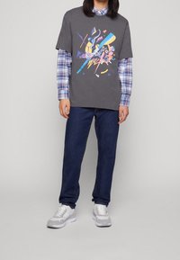 Graues Grafik-T-Shirt, das über einem blauen karierten Hemd getragen wird, kombiniert mit dunklen Jeans und grauen Sneakern; das Shirt hat ein farbenfrohes, abstraktes Design.