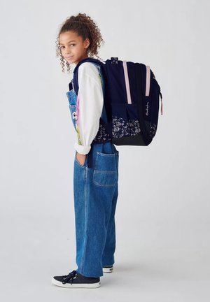 Ragazza con capelli ricci che indossa una salopette di jeans, una maglia bianca a maniche lunghe e scarpe da ginnastica, con uno zaino grande blu navy con motivi floreali.