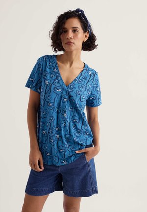 Žena s kratkom kovrčavom kosom, odjevena u plavu cvjetnu bluzu s V-izrezom, plave traper kratke hlače i odgovarajuću tamnoplavu traku za kosu, stoji s jednom rukom u džepu.