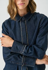 Vrouw in een donkerdenim shirt met rhinestone details langs de knopenrij en manchetten, met haar armen over haar borst gekruist.
