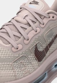 Light beige athletic shoe with textured upper, logo in dark brown, reinforced eyelets, and padded tongue with light gray accents.