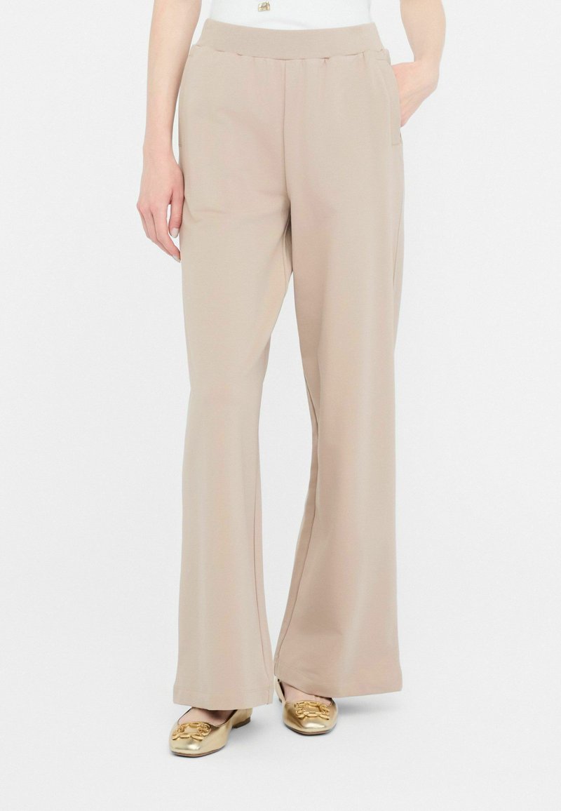 Pantaloni bej, ușori, evazați, cu un brâu elastic și buzunare laterale, asortați cu încălțăminte metalizată aurie. Textură netedă.