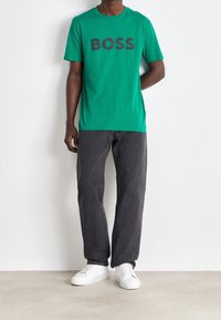 Grøn bomulds t-shirt med "BOSS" i mørkegrå skrift, parret med mørkegrå bukser og hvide sneakers.