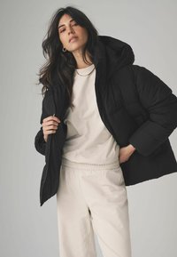 Svart puffjacka med huva, med en kort design och kviltat mönster, bärs över en beige topp och matchande ljusa byxor.