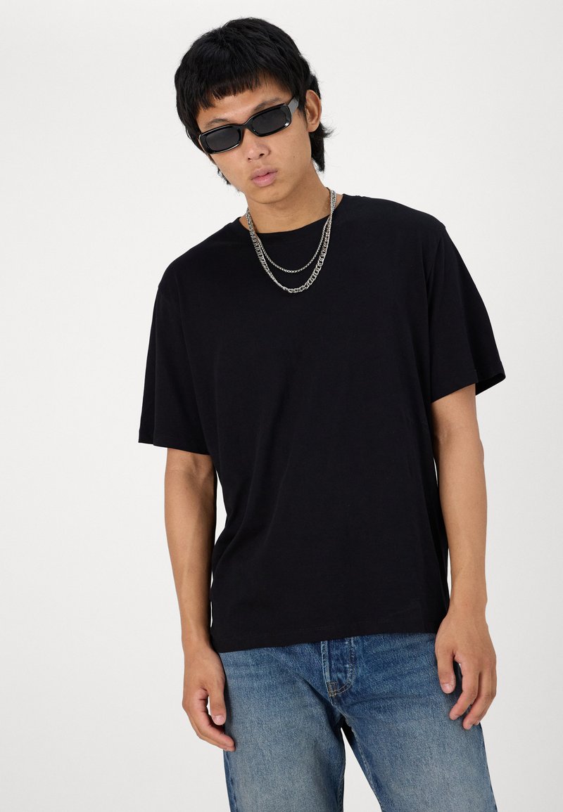 Camiseta de algodón negra de manga corta, con un corte relajado, combinada con vaqueros de mezclilla azul. Presenta accesorios de cadenas de plata superpuestos y gafas de sol negras.