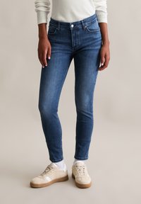 Cięte, ciemnoniebieskie dżinsy skinny z denimu, z kieszeniami z przodu, zamkiem błyskawicznym i delikatnym wyblaknięciem, w połączeniu z beżowymi trampkami i białymi skarpetkami.