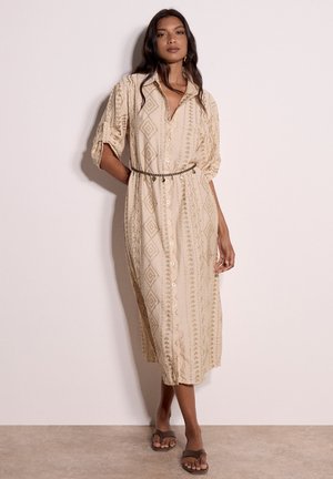 Femme portant une robe midi beige boutonnée avec des motifs géométriques, une fine ceinture et des sandales, debout contre un mur clair uni.