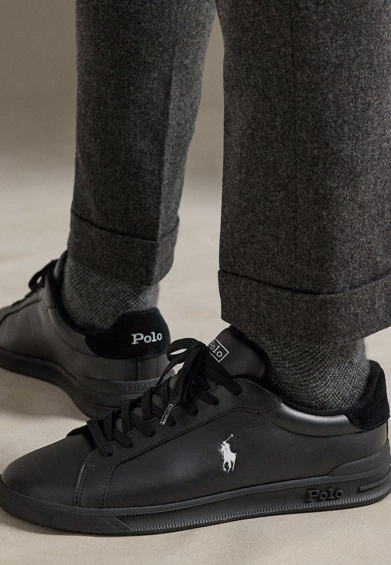 Černé kožené tenisky Polo Ralph Lauren nošené se šedými vlněnými kalhotami a vzorovanými šedými ponožkami na světlém podkladu.