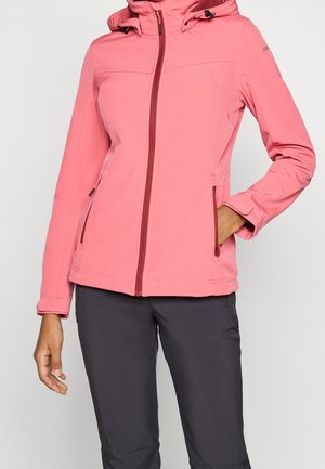 Frau trägt eine rosa Kapuzenjacke mit Reißverschluss, die Hand in der Tasche und schwarze Hose, steht vor einem schlichten hellen Hintergrund.