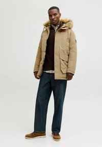 Giacca puffer khaki con cappuccio in pelliccia sintetica, due tasche frontali e chiusura con zip. Abbinata a pantaloni in denim scuri e scarpe marroni con lacci.
