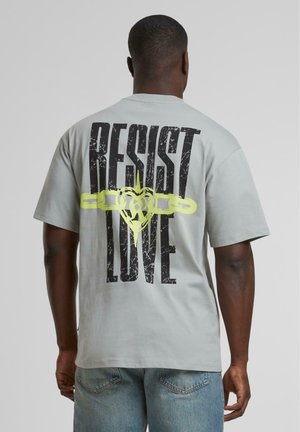 Graues T-Shirt mit kurzen Ärmeln, das mit fettem schwarzen und neongelben Grafiktext "RESIST LOVE" sowie einem herzförmigen Kettendesign auf der Rückseite gestaltet ist.