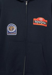Tmavomodrá mikina na zips s dvoma nášivkami s logami: kruhové logo Toyota Land Cruiser a obdĺžnikové logo Toyota s textom. Má predné vrecko.